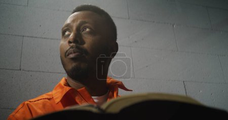 Foto de Un prisionero afroamericano con uniforme naranja se sienta en la celda de la prisión, lee la Biblia. Hombre criminal cumple pena de prisión por delito en la cárcel. Delincuente en un centro de detención o correccional. Retrato. - Imagen libre de derechos