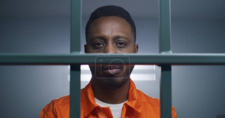 Téléchargez les photos : Afro-Américain en uniforme orange garde les mains menottées sur les barreaux de prison et regarde la caméra. Triste criminel purge une peine d'emprisonnement. Prisonnier dans un établissement correctionnel. - en image libre de droit