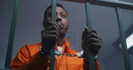 Hombre afroamericano con uniforme naranja mantiene las manos esposadas en las barras de celdas de la cárcel. Asesino deprimido cumple condena de prisión en prisión. Preso culpable en correccional o centro de detención.