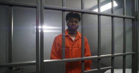 Foto de Hombre afroamericano con uniforme naranja se apoya en las barras de las celdas de la prisión y mira a la cámara. Preso deprimido cumple condena de prisión en la cárcel. Asesino en correccional o centro de detención. - Imagen libre de derechos