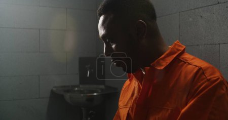 Téléchargez les photos : Un prisonnier afro-américain en uniforme orange lit la Bible en cellule, tourne des pages. Homme criminel, détenu purge une peine d'emprisonnement pour crime en prison ou dans un centre de détention. Concept de foi en Dieu. - en image libre de droit