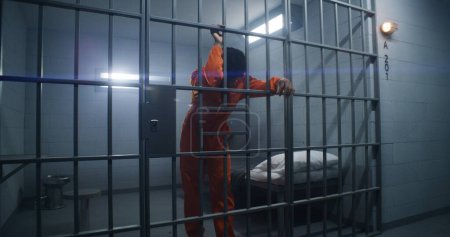 Foto de El prisionero afroamericano con uniforme naranja se apoya en las rejas de las celdas y mira a su alrededor. Gángster culpable cumple condena de prisión en correccional. Recluso sombrío en el centro de detención. - Imagen libre de derechos