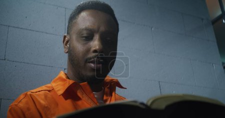 Photo for African American prisoner in orange uniform sits in prison cell, reads Bible. Male criminal serves imprisonment term for crime in jail. Offender in detention center or correctional facility. Portrait. - Royalty Free Image