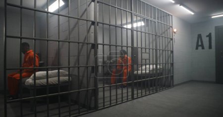 Photo pour Détenus afro-américains dans un centre de détention ou un établissement correctionnel. Homme déprimé en uniforme orange assis sur le lit de la prison et regarde la fenêtre barrée. Les prisonniers purgent une peine d'emprisonnement en cellule. - image libre de droit