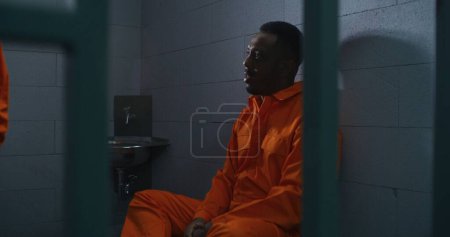 Téléchargez les photos : Un Afro-Américain est assis sur le lit et parle avec un criminel en prison. Deux prisonniers en uniforme orange dans un établissement correctionnel. Les détenus purgent une peine d'emprisonnement pour crimes dans un centre de détention. - en image libre de droit