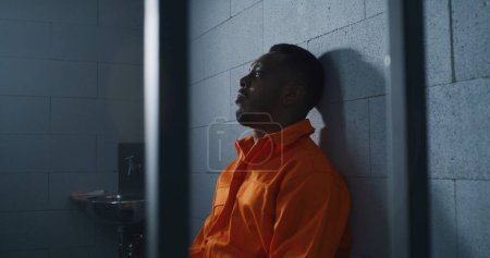 Photo for Guilty African American man in orange uniform sits on prison bed and thinks about freedom. Gloomy criminal in correctional facility or detention center. Prisoner serves imprisonment term in jail cell. - Royalty Free Image