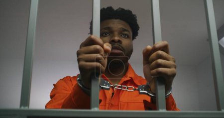 Téléchargez les photos : Un prisonnier afro-américain en uniforme orange garde les mains menottées sur les barreaux de prison. Un criminel coupable purge une peine d'emprisonnement. Triste meurtrier dans un établissement correctionnel ou un centre de détention. - en image libre de droit