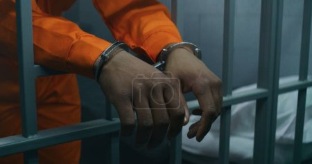 Close up shot of hands in handcuffs leaning on prison cell bars. Criminal serves imprisonment term in correctional facility or detention center. African American prisoner in orange uniform in jail.