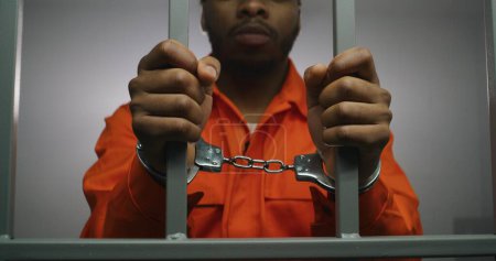 Foto de El prisionero afroamericano con uniforme naranja mantiene las manos esposadas en las barras de las celdas de la cárcel. El criminal culpable cumple condena de prisión en prisión. Asesino triste en correccional o centro de detención. - Imagen libre de derechos