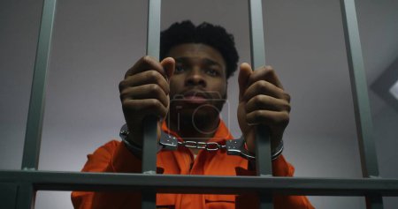 Téléchargez les photos : Un Afro-Américain en uniforme orange garde les mains menottées sur les barreaux de prison. Un meurtrier déprimé purge une peine d'emprisonnement. Prisonnier coupable dans un établissement correctionnel ou un centre de détention. - en image libre de droit