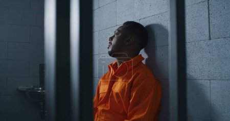 Téléchargez les photos : Coupable homme afro-américain en uniforme orange assis sur le lit de prison et pense à la liberté. Criminel sombre dans un établissement correctionnel ou un centre de détention. Un prisonnier purge une peine d'emprisonnement en cellule. - en image libre de droit