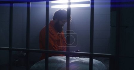 Photo for Believing male prisoner in orange uniform kneels near the bed, prays to God in prison cell holding Bible. Criminal serves imprisonment term for crime in jail or detention center. Faith in God concept. - Royalty Free Image