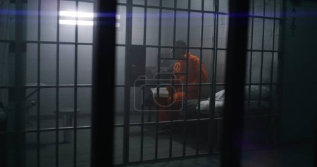 Téléchargez les photos : Un prisonnier afro-américain en uniforme orange est assis sur le lit, lit la Bible en cellule de prison. Homme criminel purge une peine d'emprisonnement pour crime en prison ou en centre de détention. La foi en Dieu. Vue à travers les barres. - en image libre de droit