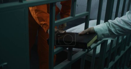 Foto de El oficial de la prisión le da la Biblia al prisionero en uniforme naranja en la celda de la prisión. Criminal cumple pena de prisión por crimen en la cárcel. Centro de detención o correccional. Vista a través de barras de metal. - Imagen libre de derechos