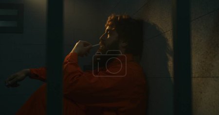 Téléchargez les photos : Un homme prisonnier déprimé en uniforme orange est assis sur le lit, fume une cigarette dans une cellule de prison. Criminel, le détenu purge une peine d'emprisonnement pour crime en prison. Centre de détention ou établissement correctionnel. - en image libre de droit