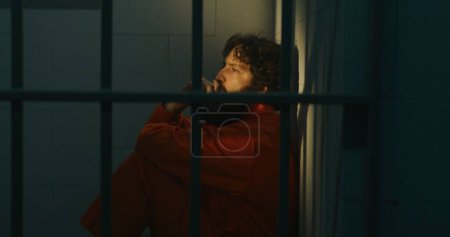 Téléchargez les photos : Un prisonnier pensif en uniforme orange s'assoit sur son lit, fume une cigarette dans sa cellule. Le criminel purge une peine d'emprisonnement pour crime en prison. Le soleil brille par la fenêtre barrée. Centre de détention. Portrait. - en image libre de droit