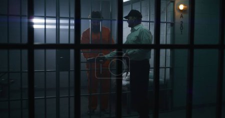 Photo for Prison officer gives Bible to male prisoner in orange uniform in prison cell. Criminal serves imprisonment term for crime in jail. Detention center or correctional facility. View through metal bars. - Royalty Free Image