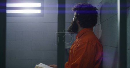 Photo for Male prisoner in orange uniform sits on the bed, reads Bible, looks at barred window in prison cell. Criminal serves imprisonment term for crime in jail. Detention center or correctional facility. - Royalty Free Image