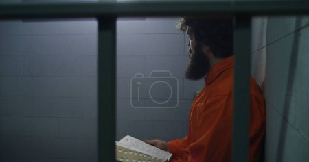 Photo for Male prisoner in orange uniform sits on the bed, reads Bible, looks at barred window in prison cell. Criminal serves imprisonment term for crime in jail. Detention center or correctional facility. - Royalty Free Image