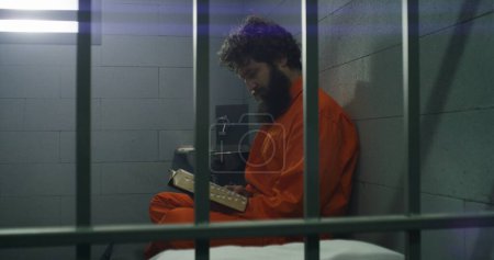 Téléchargez les photos : Un prisonnier en uniforme orange est assis sur le lit, lit la Bible, regarde une fenêtre barrée dans une cellule de prison. Le criminel purge une peine d'emprisonnement pour crime en prison. Centre de détention ou établissement correctionnel. - en image libre de droit
