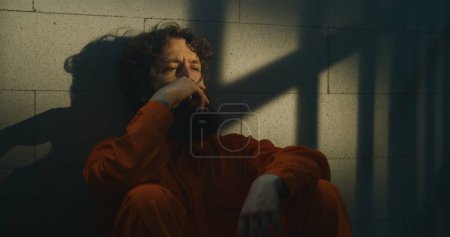 Téléchargez les photos : Prisonnier déprimé en uniforme orange assis sur le lit, fume une cigarette dans une cellule de prison. Le criminel purge une peine d'emprisonnement pour crime en prison. Le soleil brille par la fenêtre barrée. Centre de détention. Portrait. - en image libre de droit
