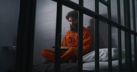 Photo for Male prisoner in orange uniform sits on bed, reads Bible, prays, looks at barred window in prison cell. Inmate serves imprisonment term for crime in jail. Correctional facility. Faith in God concept. - Royalty Free Image