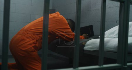 Foto de Religioso prisionero afroamericano en uniforme naranja se arrodilla cerca de la cama, reza a Dios en la celda de la prisión sosteniendo la Biblia. El delincuente masculino cumple condena de prisión en la cárcel o centro de detención. Fe en Dios. - Imagen libre de derechos