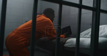 Foto de Religioso prisionero afroamericano en uniforme naranja se arrodilla cerca de la cama, reza a Dios en la celda de la prisión sosteniendo la Biblia. El delincuente masculino cumple condena de prisión en la cárcel o centro de detención. Fe en Dios. - Imagen libre de derechos