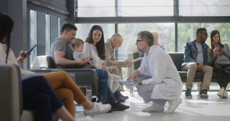 Photo for Diverse people sit on couches in clinic lobby area, wait for doctors appointment. Doctor talks to family with little child about medical test results. Waiting room in modern medical center. Healthcare - Royalty Free Image