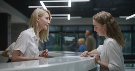 Photo for Female administrator talks with patient at reception desk in clinic lobby area. Woman consults person, helps fill out papers in hospital. Medical staff works in modern medical center. Health care. - Royalty Free Image