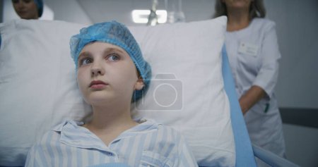 Photo for Young girl sits on gurney and looks around. Professional medics walk down medical center hallway, push gurney with patient to hospital room after surgery. Medical personnel at work in modern clinic. - Royalty Free Image