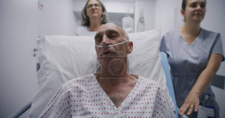 Photo for Old patient lies on gurney with breathing tube, talks with female doctor and nurse. Medics transport senior man on surgery to operating room down hospital hallway. Medical personnel at work in clinic. - Royalty Free Image