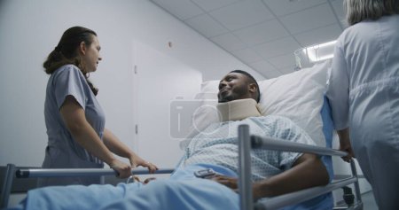 Téléchargez les photos : Les infirmières transportent les patients afro-américains dans le couloir de la clinique. Homme d'âge moyen se trouve sur le brancard dans l'attelle du cou et avec oxymètre de pouls, parle avec l'infirmière de sa santé. Personnel médical au travail. - en image libre de droit