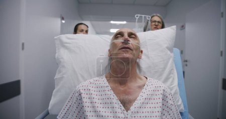 Photo for Senior patient with breathing tube sleeps on gurney before operation. Mature doctor with young nurse transports old man to operation room down medical center corridor. Medical staff in modern clinic. - Royalty Free Image