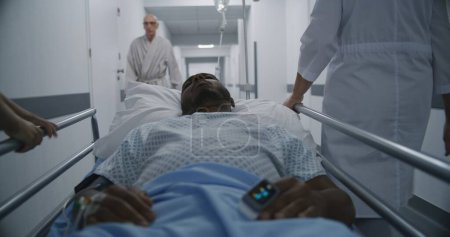 Photo for African American patient sleeps on gurney with venous catheter and pulse oximeter on hands after serious operation. Doctor and nurse transport middle aged man to hospital room. Medics save lifes. - Royalty Free Image