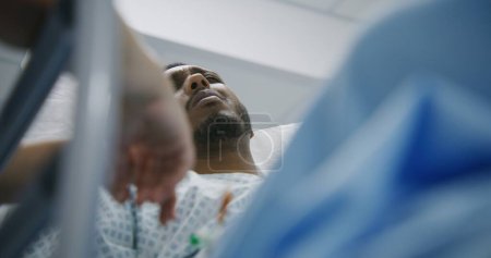 Photo for African American man lies on stretcher and talks with doctors before surgery. Physician stands near gurney and calms patient in medical center. Medical staff work in emergency department. Close up. - Royalty Free Image