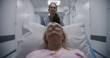 Téléchargez les photos : Une patiente âgée dort sur une civière avec un tube respiratoire après l'opération. Une jeune infirmière transporte une femme âgée dans une chambre d'hôpital au bout du couloir. Personnel médical au travail dans un centre médical. - en image libre de droit