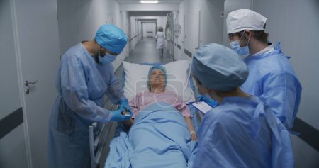 Foto de Médicos, enfermeras y paramédicos examinan a una anciana enferma en el corredor de las instalaciones médicas. El médico comprueba el oxímetro de pulso en la mano del paciente. El personal médico trabaja en urgencias. Acercar. - Imagen libre de derechos