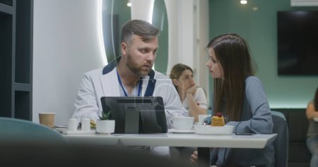 Photo for Professional doctor shows medical tests results to patient on digital tablet computer in hospital cafe and discusses it. Female medic eats dinner in the background. Medical staff in hospital canteen. - Royalty Free Image