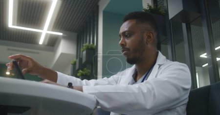 Téléchargez les photos : Un médecin afro-américain est assis dans un café de clinique. Médecin professionnel travaille sur tablette numérique, regarde les résultats des tests médicaux des patients. Le personnel médical a une pause à la cafétéria de l'hôpital ou du centre médical. - en image libre de droit