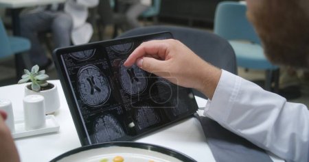 Photo for Doctor watches image of MRI or CT scan using digital tablet in hospital cafe. Medic eats his dinner, analyzes brain scanning results of patient. Medical staff have meal in clinic cafeteria. Close up. - Royalty Free Image