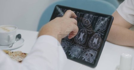 Photo for Professional doctor looks at image of MRI or CT scan on digital tablet computer in hospital cafe. Medic examines brain scanning results of patient. Medical staff have brake in clinic cafeteria. - Royalty Free Image