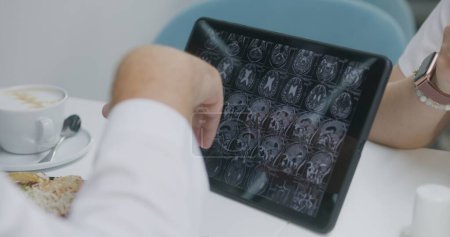 Foto de El médico profesional examina la imagen de la resonancia magnética o la tomografía computarizada en la tableta digital en la cafetería del hospital. El médico examina los resultados de la exploración cerebral del paciente. El personal médico tiene freno en la cafetería de la clínica. - Imagen libre de derechos