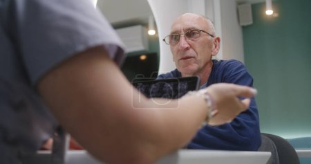 Photo for Elderly patient sits in clinic cafe with doctor. Professional medic discusses medical diagnostic or tests results with man. Digital tablet with MRI scan image. Hospital or medical center dining room. - Royalty Free Image