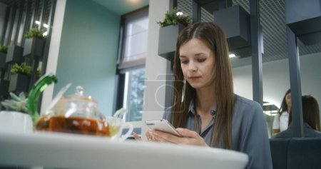 Photo for Woman sits at the table in modern clinic cafe after appointment with doctor, uses phone. Cafeteria worker brings meal to female visitor. Patient of medical center or hospital having dinner in canteen. - Royalty Free Image