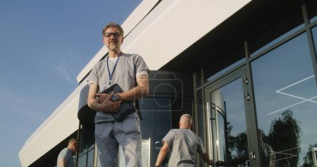 Photo for Mature doctor with tablet in hands stands with patient at clinic entrance, discuss treatment. Healthcare specialist says goodbye to client after appointment, then looks at camera. Medic works outside. - Royalty Free Image