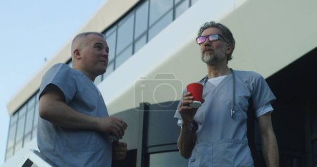 Photo for Two doctors in uniform stand near medical center entrance in the evening. Professional medics drink coffee during break and talk. Medical staff of hospital or modern clinic. Lifestyle and healthcare. - Royalty Free Image
