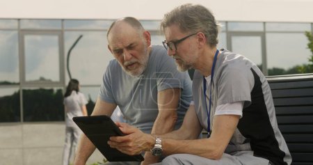 Photo for Mature doctor with tablet in hands sits with elderly patient on bench and consults him. Professional in medical uniform makes diagnosis to client using digital tablet. Medical staff work outdoor. - Royalty Free Image