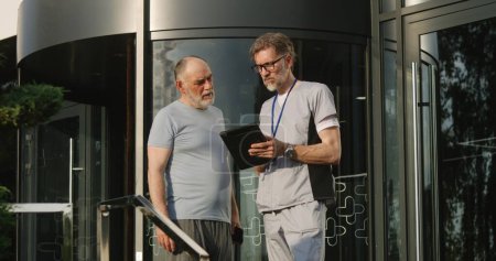 Photo for Mature physician with digital tablet in hands stands with aged patient at clinic entrance and discuss his treatment. Health care specialist consults and supports elderly client. Medic works outside. - Royalty Free Image