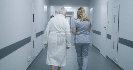 Female doctor, nurse with digital tablet walks along the clinic corridor with elderly woman, helps patient to get to hospital ward after procedures. Medical staff, patients in medical center hallway.